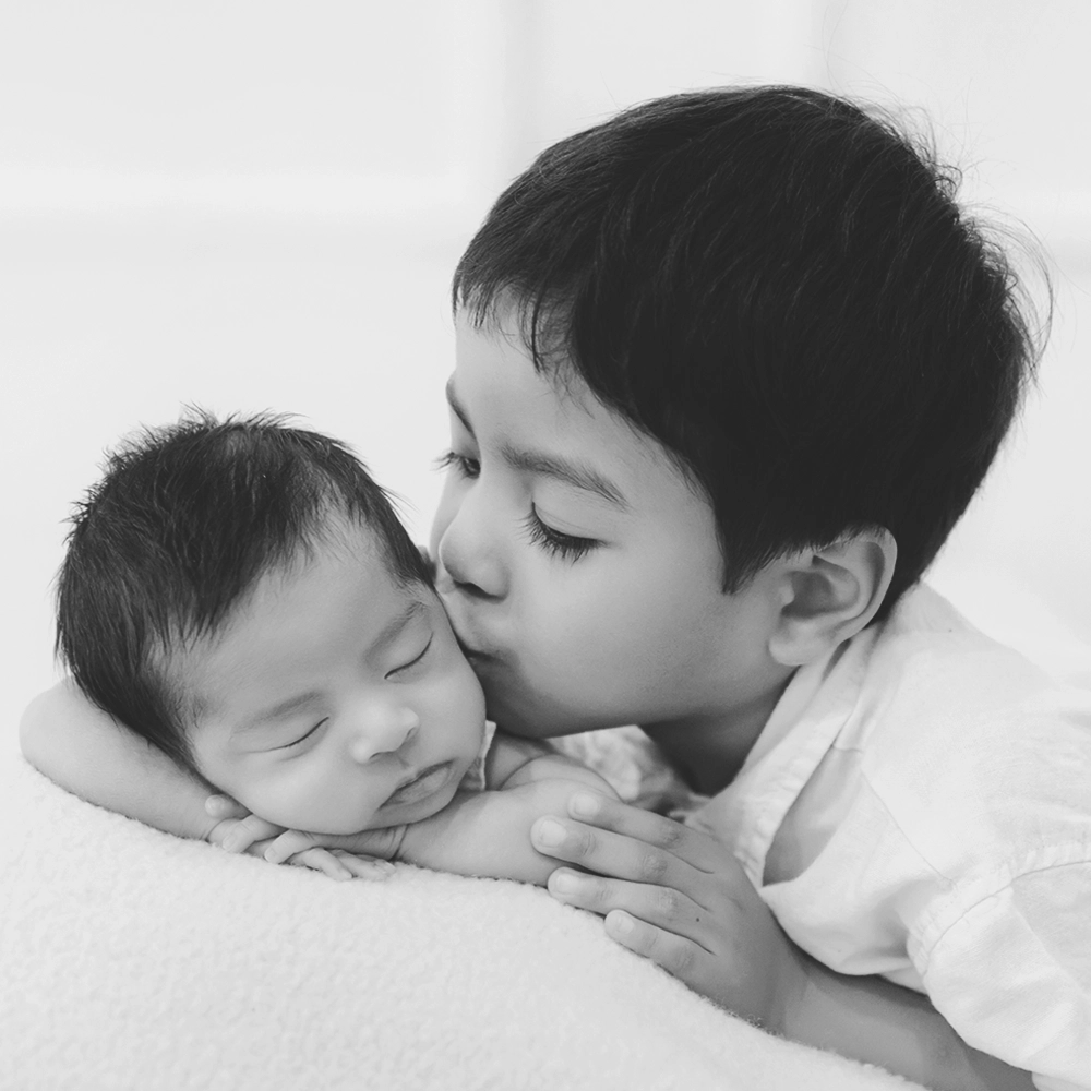 Photo d'un bébé faisant une pose et son grand frère qui lui fait un bisous de côté lors d'un shooting photo avec Tiana Photographe Toulouse